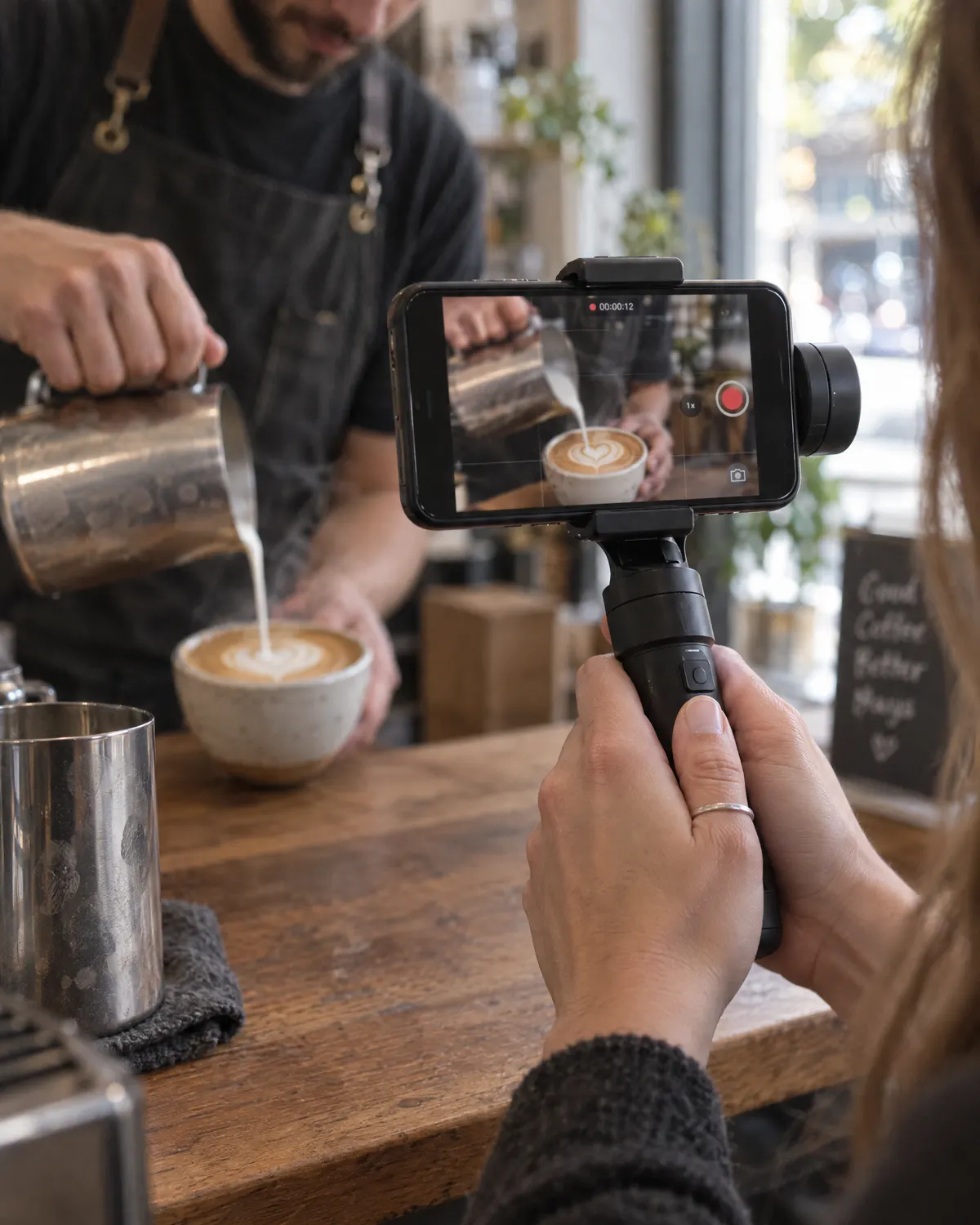 Hands holding a phone on a gimbal, filming a barista pouring latte art